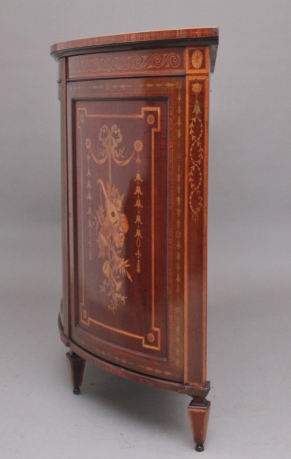 A 19th-century bow-fronted mahogany side cabinet featuring detailed satinwood marquetry panels with classical figures and swags on turned tapered legs. View VIII