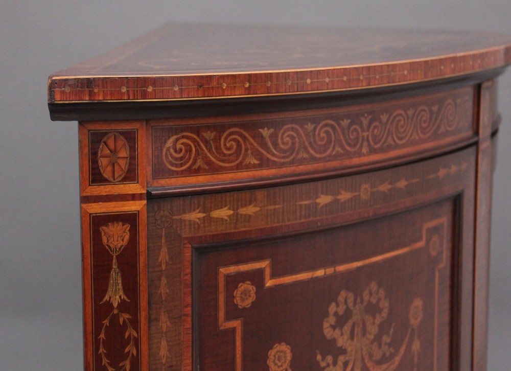A 19th-century bow-fronted mahogany side cabinet featuring detailed satinwood marquetry panels with classical figures and swags on turned tapered legs. View X