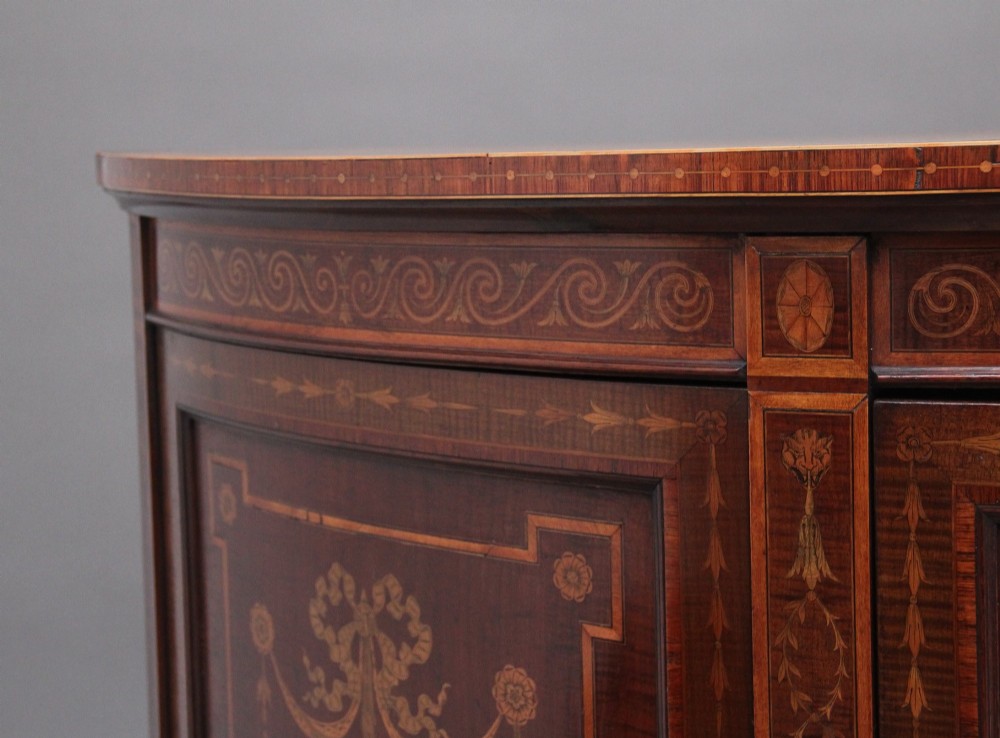 A 19th-century bow-fronted mahogany side cabinet featuring detailed satinwood marquetry panels with classical figures and swags on turned tapered legs. View XI