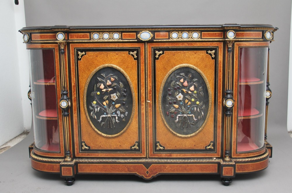 A 19th-century Victorian amboyna and ebonised credenza featuring curved glass side cabinets, gilt-metal mounts, porcelain plaque frieze details, and central oval panels inlaid with stone floral motifs.