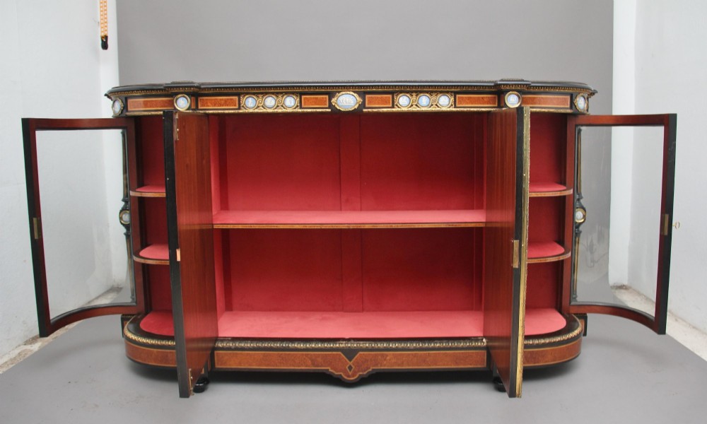 A 19th-century Victorian amboyna and ebonised credenza featuring curved glass side cabinets, gilt-metal mounts, porcelain plaque frieze details, and central oval panels inlaid with stone floral motifs. View II