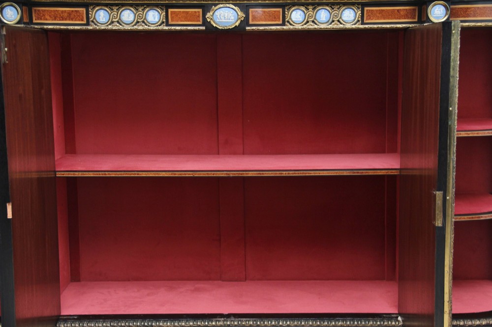 A 19th-century Victorian amboyna and ebonised credenza featuring curved glass side cabinets, gilt-metal mounts, porcelain plaque frieze details, and central oval panels inlaid with stone floral motifs. View IV