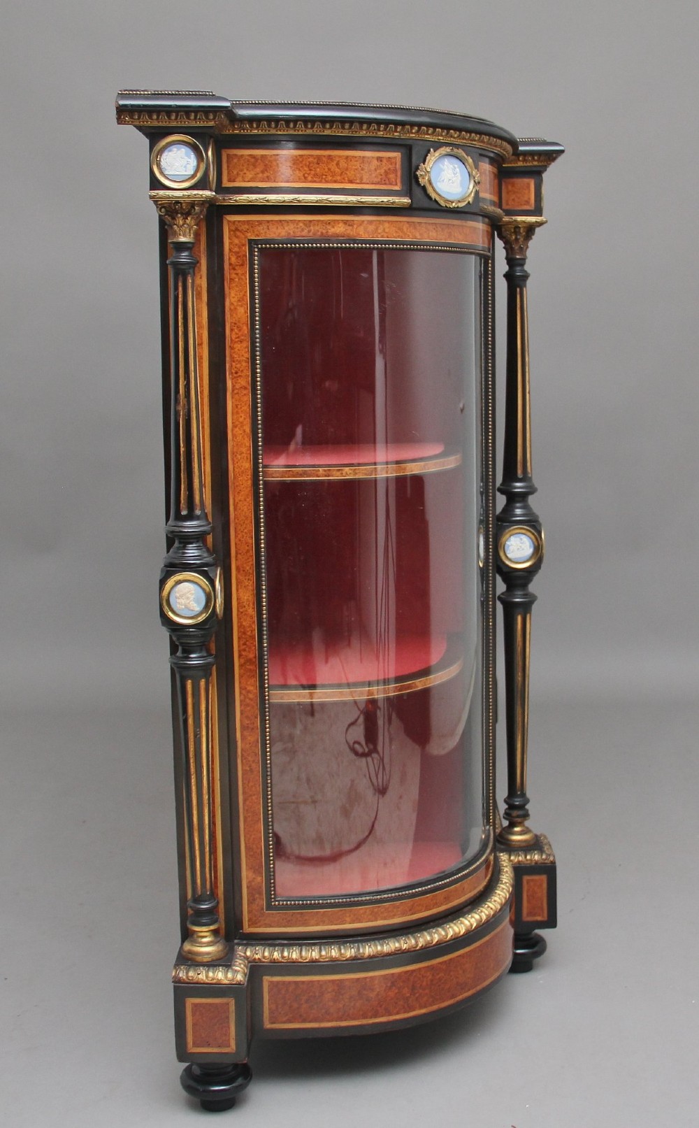 A 19th-century Victorian amboyna and ebonised credenza featuring curved glass side cabinets, gilt-metal mounts, porcelain plaque frieze details, and central oval panels inlaid with stone floral motifs. View V