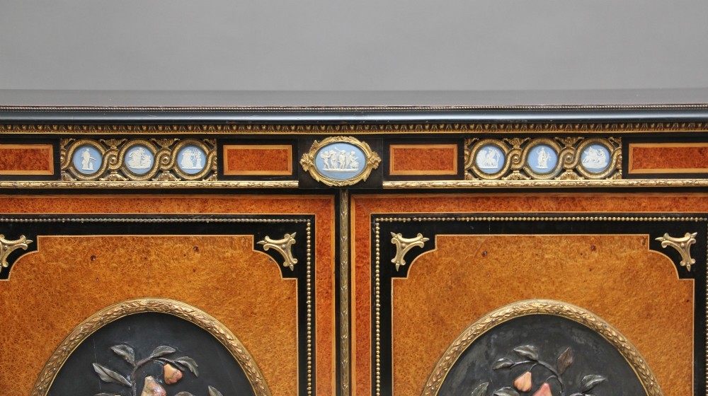 A 19th-century Victorian amboyna and ebonised credenza featuring curved glass side cabinets, gilt-metal mounts, porcelain plaque frieze details, and central oval panels inlaid with stone floral motifs. View XIV