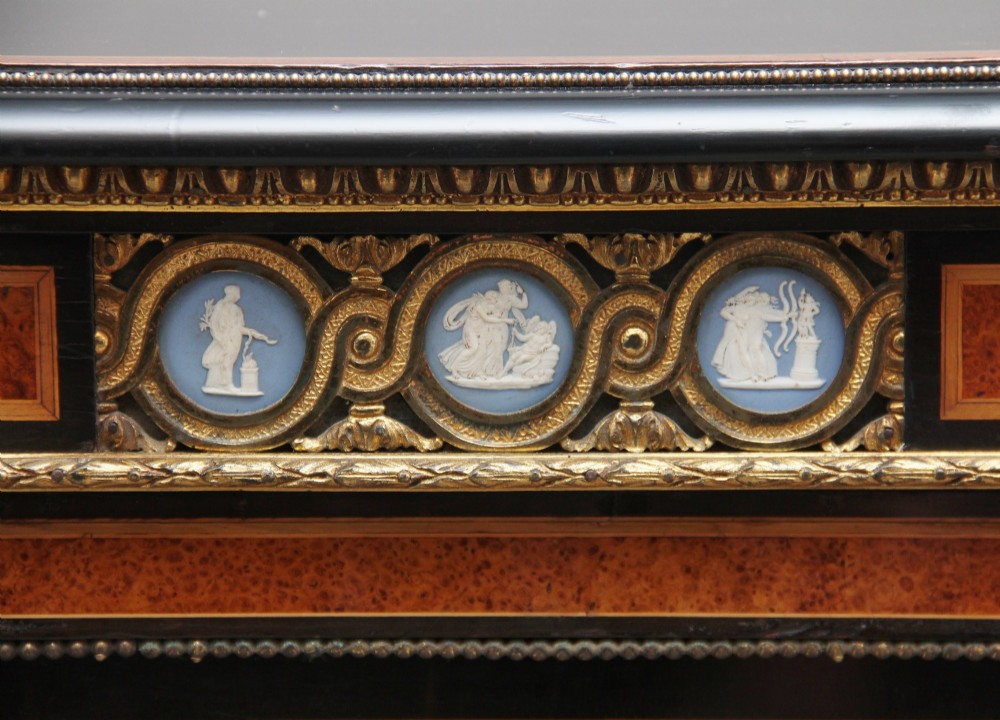 A 19th-century Victorian amboyna and ebonised credenza featuring curved glass side cabinets, gilt-metal mounts, porcelain plaque frieze details, and central oval panels inlaid with stone floral motifs. View XV