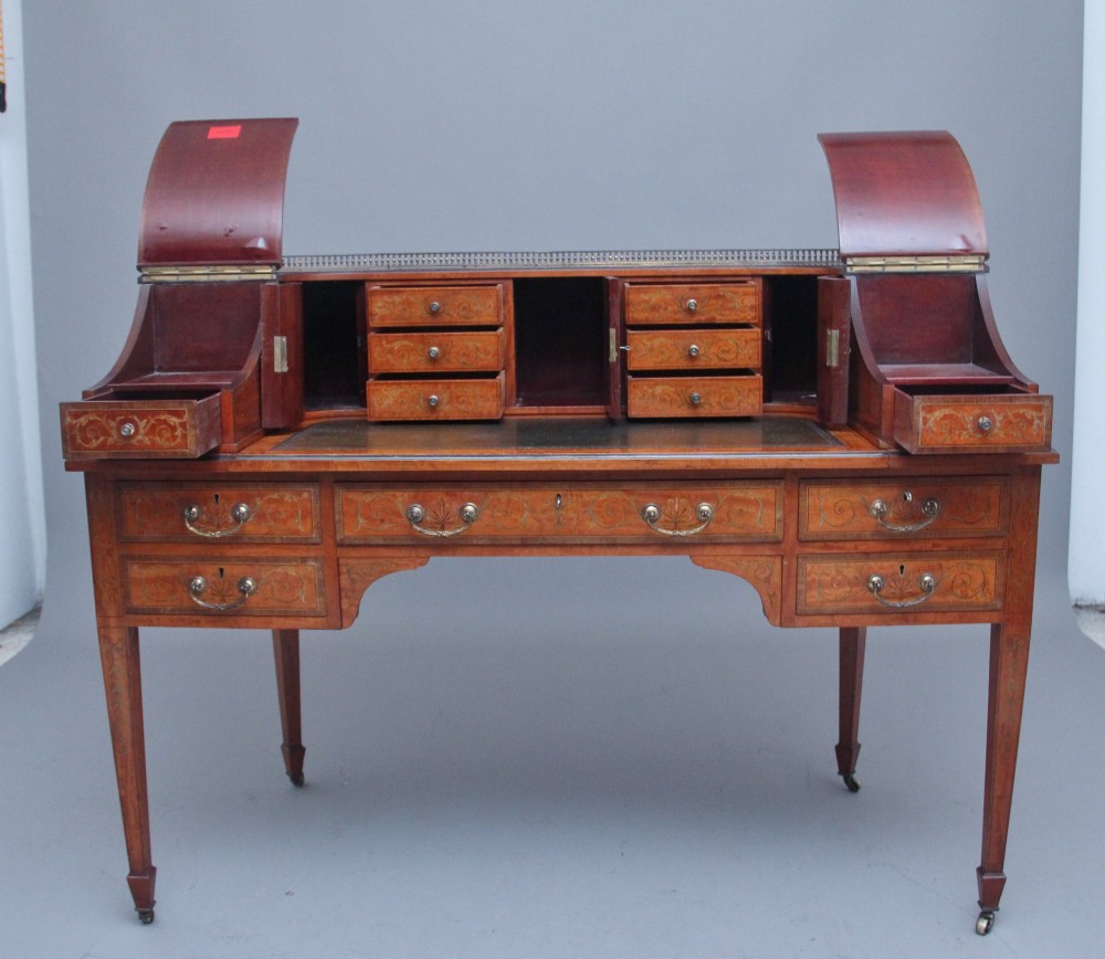 A 19th-century satinwood Carlton House desk featuring floral marquetry inlay, multiple small drawers, a central writing surface, a brass gallery rail, and square tapered legs on castors. View II