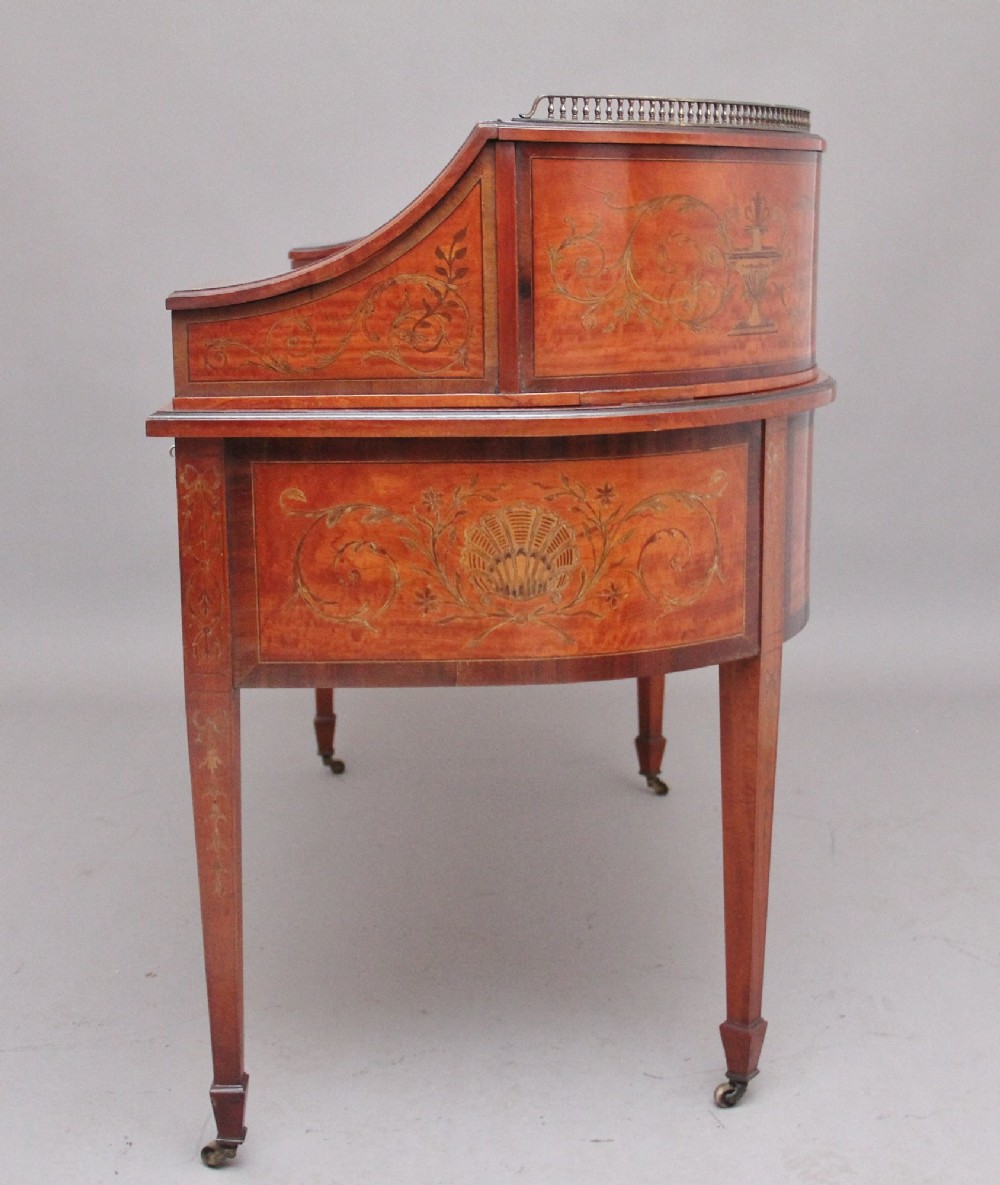 A 19th-century satinwood Carlton House desk featuring floral marquetry inlay, multiple small drawers, a central writing surface, a brass gallery rail, and square tapered legs on castors. View XII