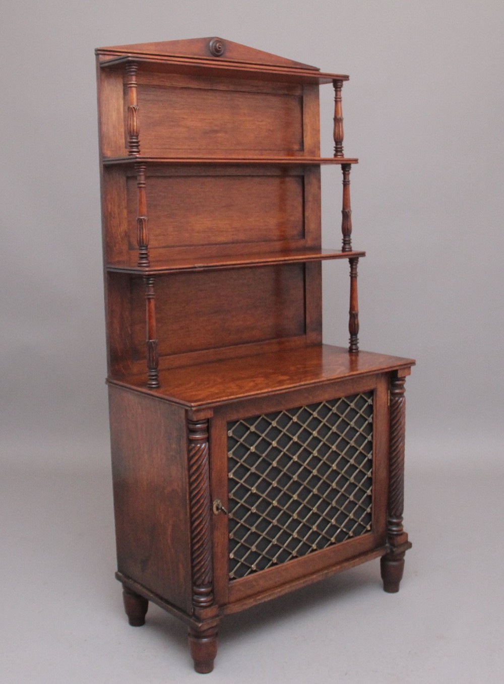 An early nineteenth-century oak bookcase featuring three open upper shelves, a pediment top, and a lower cabinet with a brass wire mesh door flanked by rope-twist columns. View II