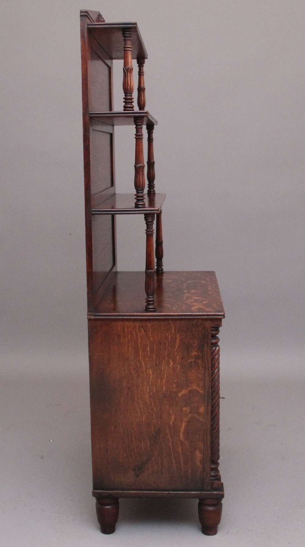 An early nineteenth-century oak bookcase featuring three open upper shelves, a pediment top, and a lower cabinet with a brass wire mesh door flanked by rope-twist columns. View VI