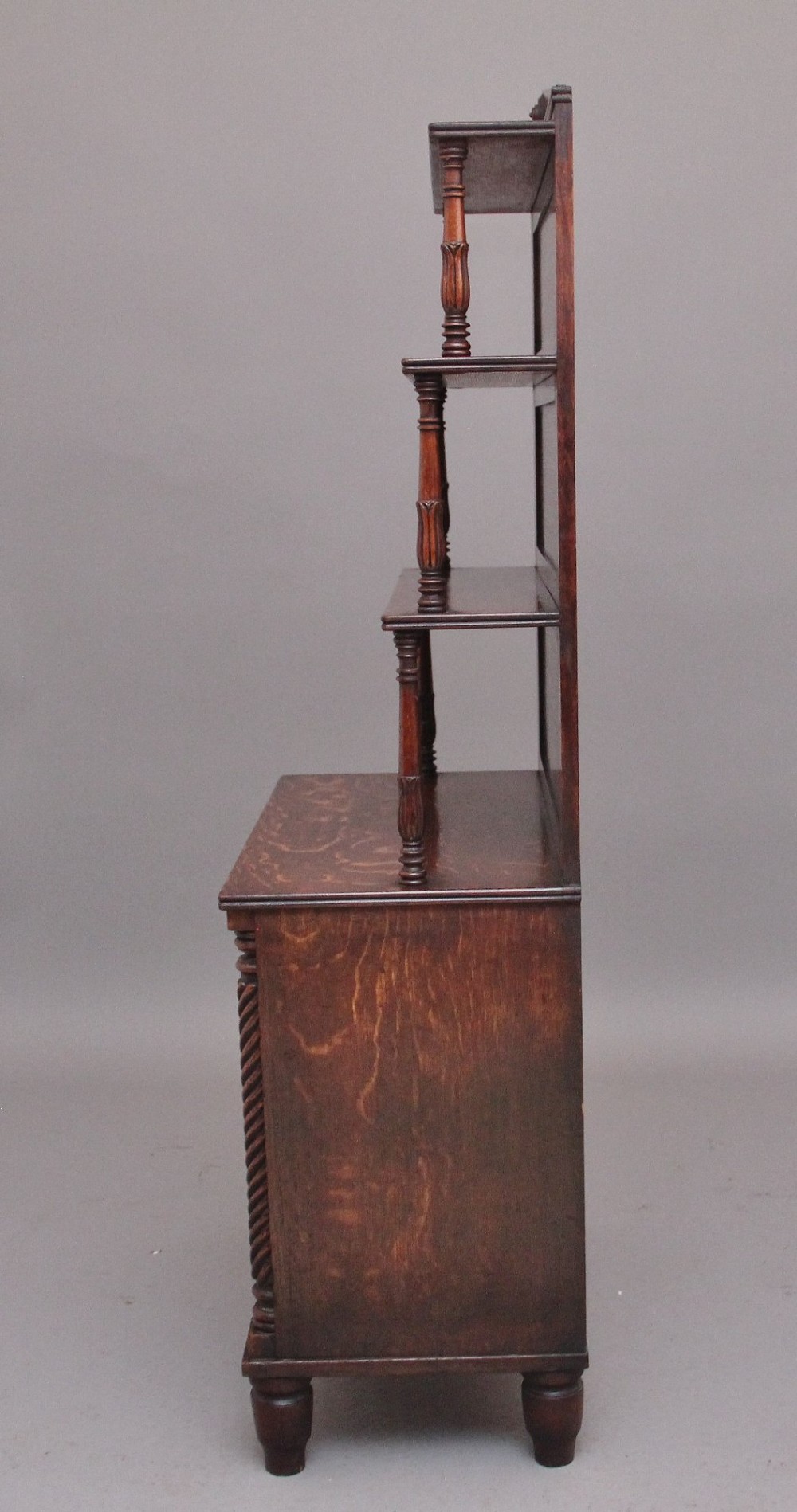 An early nineteenth-century oak bookcase featuring three open upper shelves, a pediment top, and a lower cabinet with a brass wire mesh door flanked by rope-twist columns. View IX
