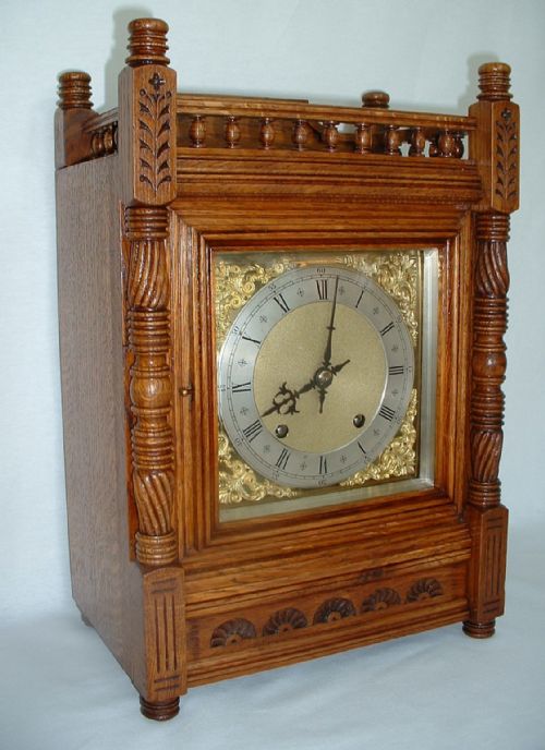 fig 1. An antique carved oak bracket clock featuring a brass dial with Roman numerals, side columns, and decorative balustrade top gallery.