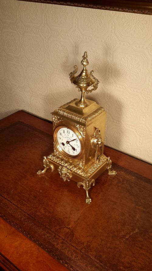 fig 1. An antique French brass mantel clock with an ornamental urn finial, intricate foliate casing, and a white enamel dial.
