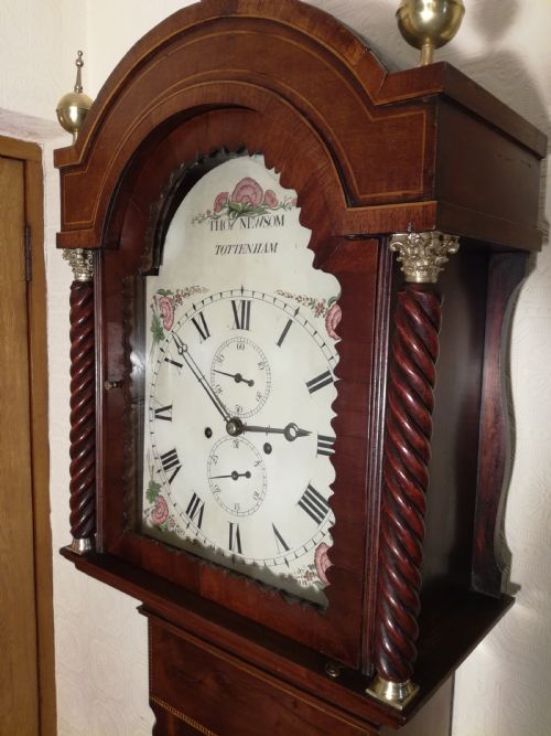fig 1. A tall Regency flame mahogany longcase clock featuring brass ball finials, spiral twisted columns flanking the hood, and an arched painted dial with Roman numerals.