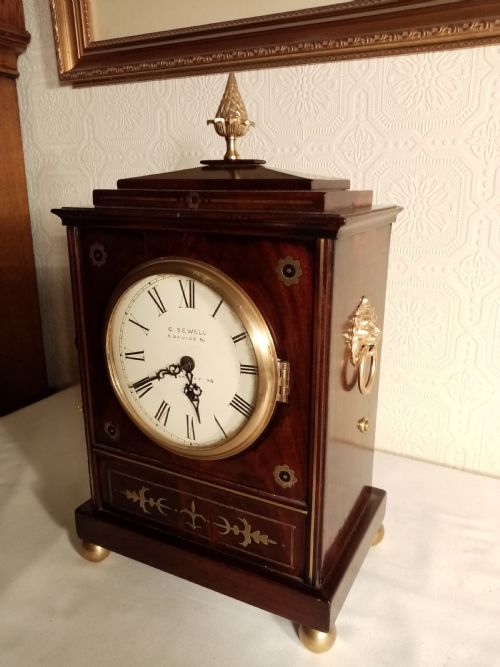 fig 1. A Regency period dark mahogany library clock featuring brass inlays, a round white dial with Roman numerals, brass ball feet, and a flame finial.