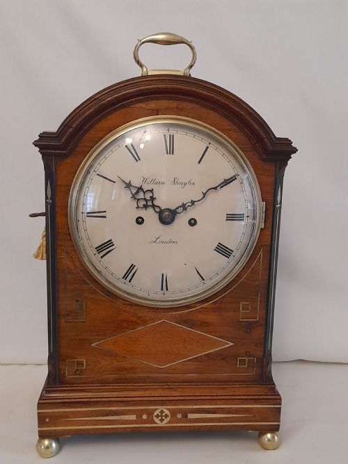 fig 1. A Regency period rosewood bracket clock featuring geometric brass inlay, arched top with brass handle, gothic side frets, circular dial, and brass ball feet.