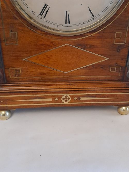 fig 2. A Regency period rosewood bracket clock featuring geometric brass inlay, arched top with brass handle, gothic side frets, circular dial, and brass ball feet.
