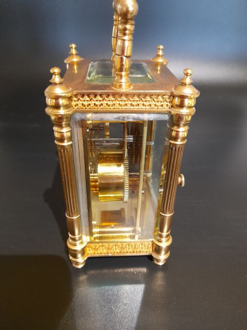 fig 2. A gilded brass carriage clock featuring clear glass side panels, turned corner columns, a folding top handle, and a white enamel dial with Arabic numerals.