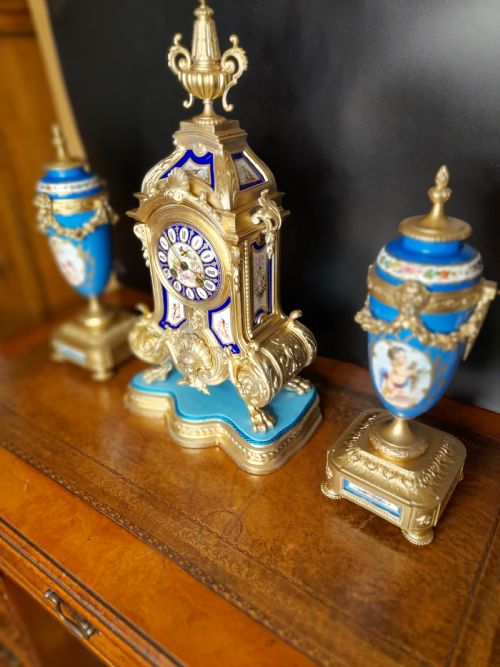 fig 1. A French gilt bronze clock garniture featuring a central mantel clock with hand-painted blue porcelain panels flanked by two matching ornamental urns.