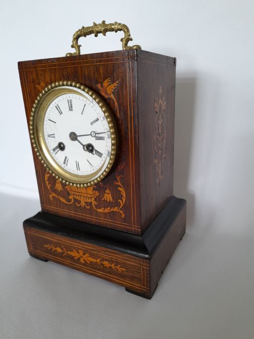 fig 1. An antique French officers clock with a dark wood inlaid rectangular case, a decorative brass carrying handle, and a white enamel clock face with Roman numerals.