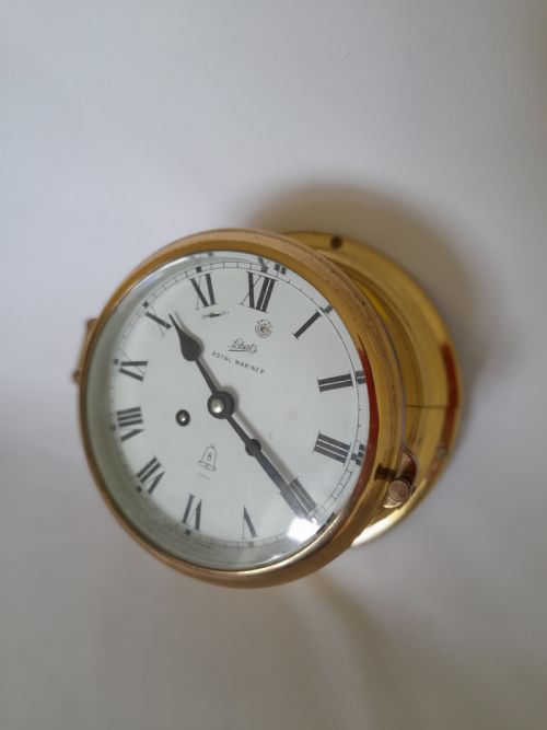 fig 1. A circular brass Schatz Royal Mariner maritime wall clock with a white face, black Roman numerals, and a hinged glass front.