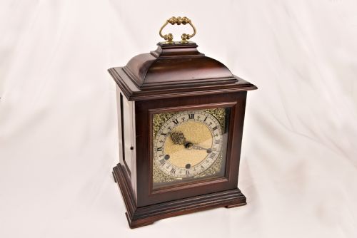fig 2. A dark mahogany bracket clock featuring a brass carrying handle on top, side glass panels, and an ornate brass dial with a silvered chapter ring.