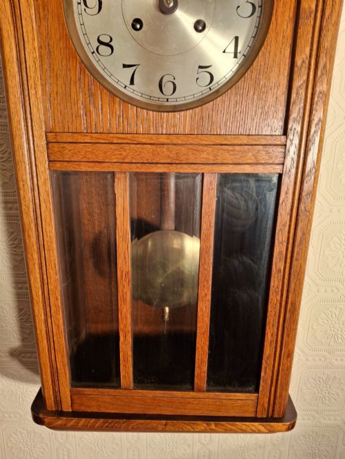 fig 1. An Arts and Crafts period golden oak wall clock featuring a circular metal dial with Arabic numerals and a lower glazed window displaying a brass pendulum.