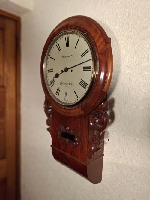 fig 1. An antique mahogany drop-dial wall clock featuring a circular white dial with Roman numerals above a carved wooden lower trunk.