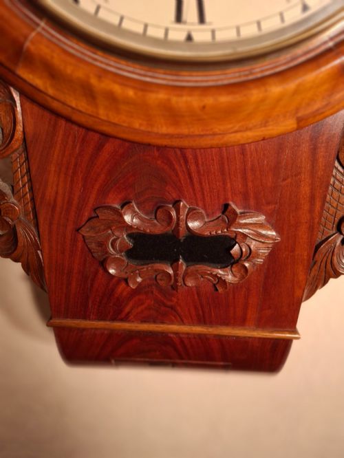 fig 2. An antique mahogany drop-dial wall clock featuring a circular white dial with Roman numerals above a carved wooden lower trunk.