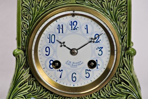 fig 1. An olive green glazed ceramic mantel clock featuring relief acanthus foliage details, a white dial with blue Arabic numerals, mounted on a dark wooden base.
