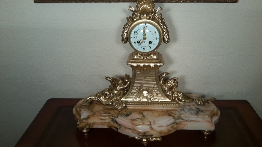 An antique French gilt metal mantel clock featuring foliage motifs and an enamel dial, mounted upon a contoured pink and beige marble base.