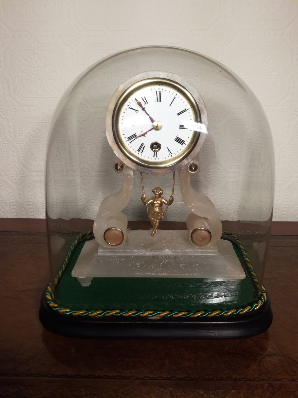 An antique French alabaster mantel clock under a clear glass dome, featuring a circular white dial above a gilt cherub figure on a swing pendulum.