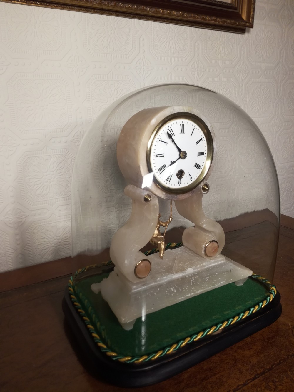 An antique French alabaster mantel clock under a clear glass dome, featuring a circular white dial above a gilt cherub figure on a swing pendulum. View II