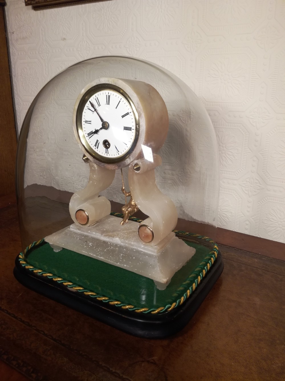 An antique French alabaster mantel clock under a clear glass dome, featuring a circular white dial above a gilt cherub figure on a swing pendulum. View III