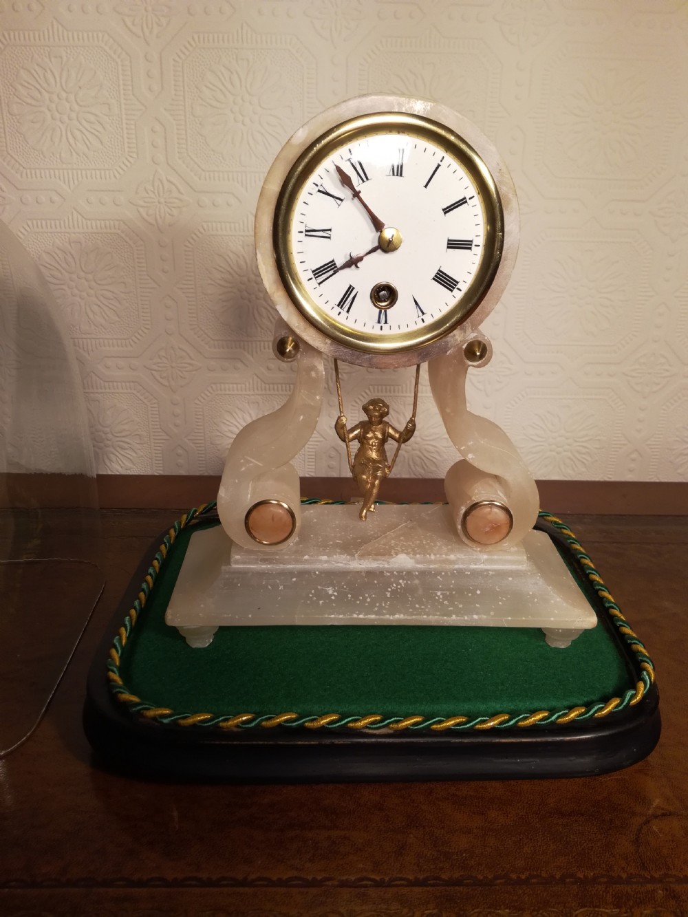 An antique French alabaster mantel clock under a clear glass dome, featuring a circular white dial above a gilt cherub figure on a swing pendulum. View V