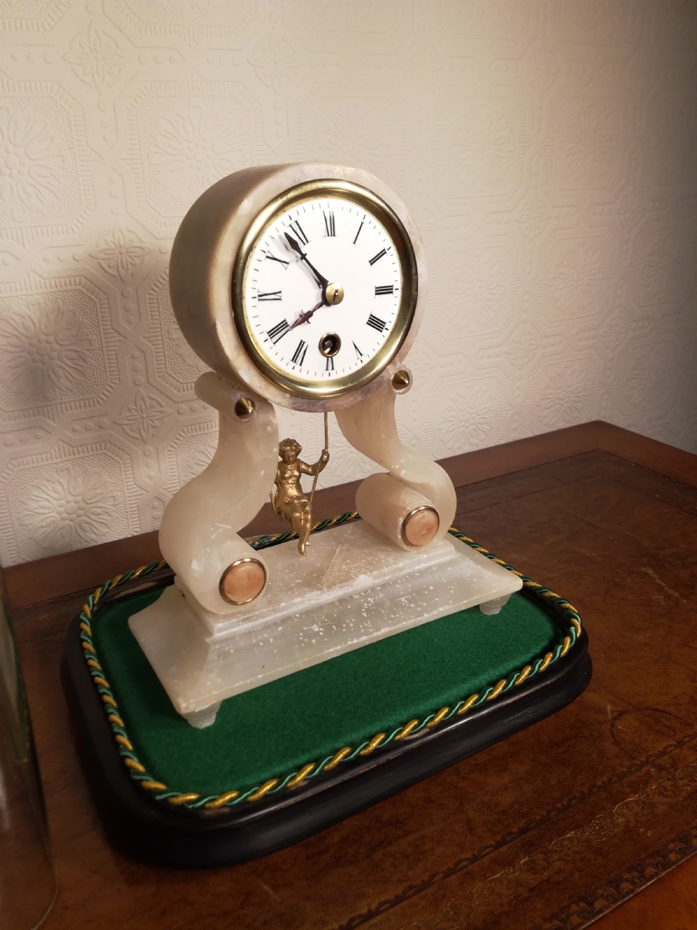 An antique French alabaster mantel clock under a clear glass dome, featuring a circular white dial above a gilt cherub figure on a swing pendulum. View VI