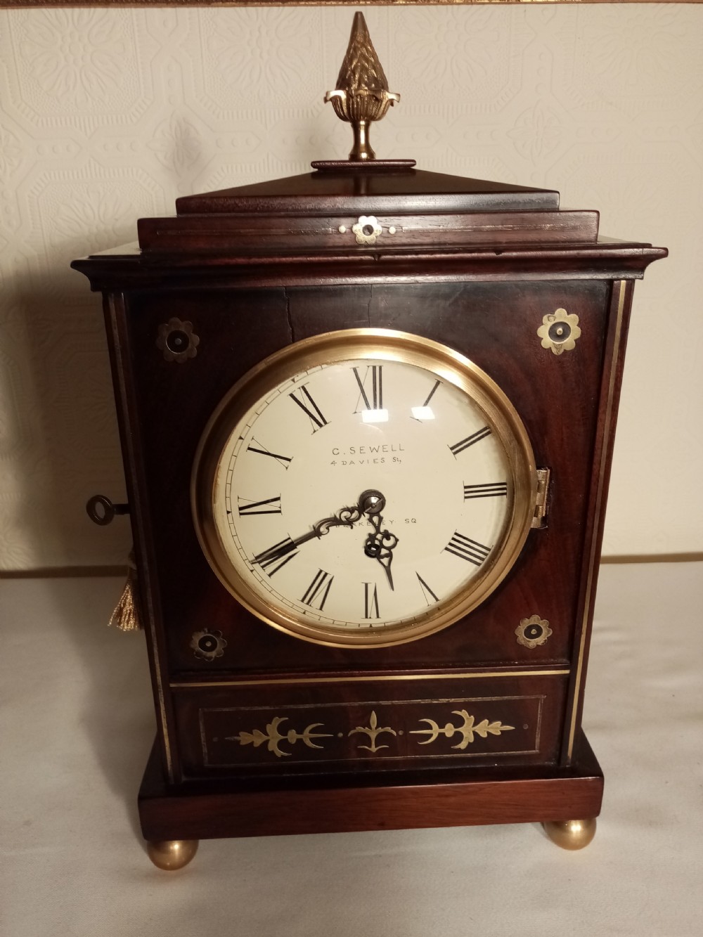 A Regency period dark mahogany library clock featuring brass inlays, a round white dial with Roman numerals, brass ball feet, and a flame finial.
