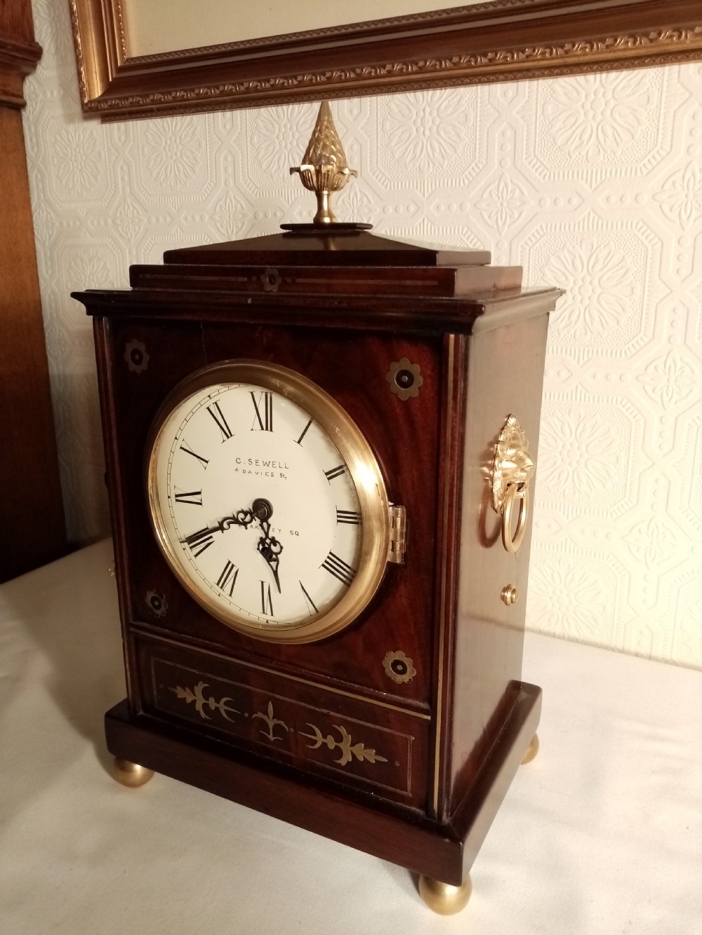 A Regency period dark mahogany library clock featuring brass inlays, a round white dial with Roman numerals, brass ball feet, and a flame finial. View II