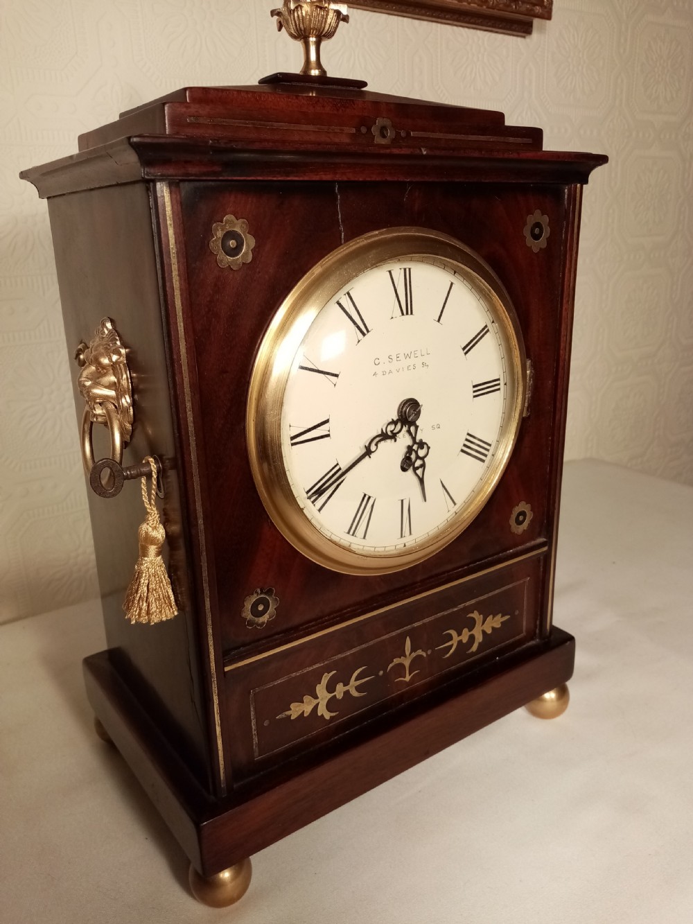 A Regency period dark mahogany library clock featuring brass inlays, a round white dial with Roman numerals, brass ball feet, and a flame finial. View III