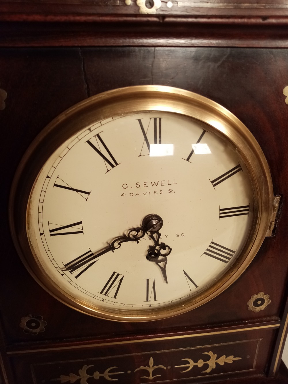 A Regency period dark mahogany library clock featuring brass inlays, a round white dial with Roman numerals, brass ball feet, and a flame finial. View V