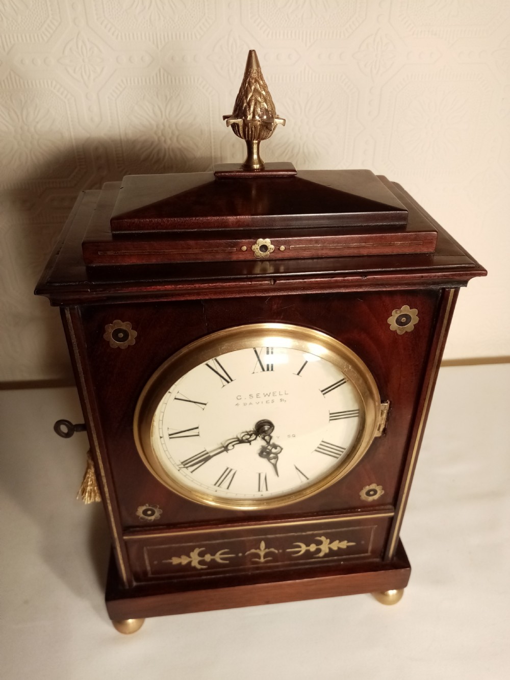A Regency period dark mahogany library clock featuring brass inlays, a round white dial with Roman numerals, brass ball feet, and a flame finial. View VI