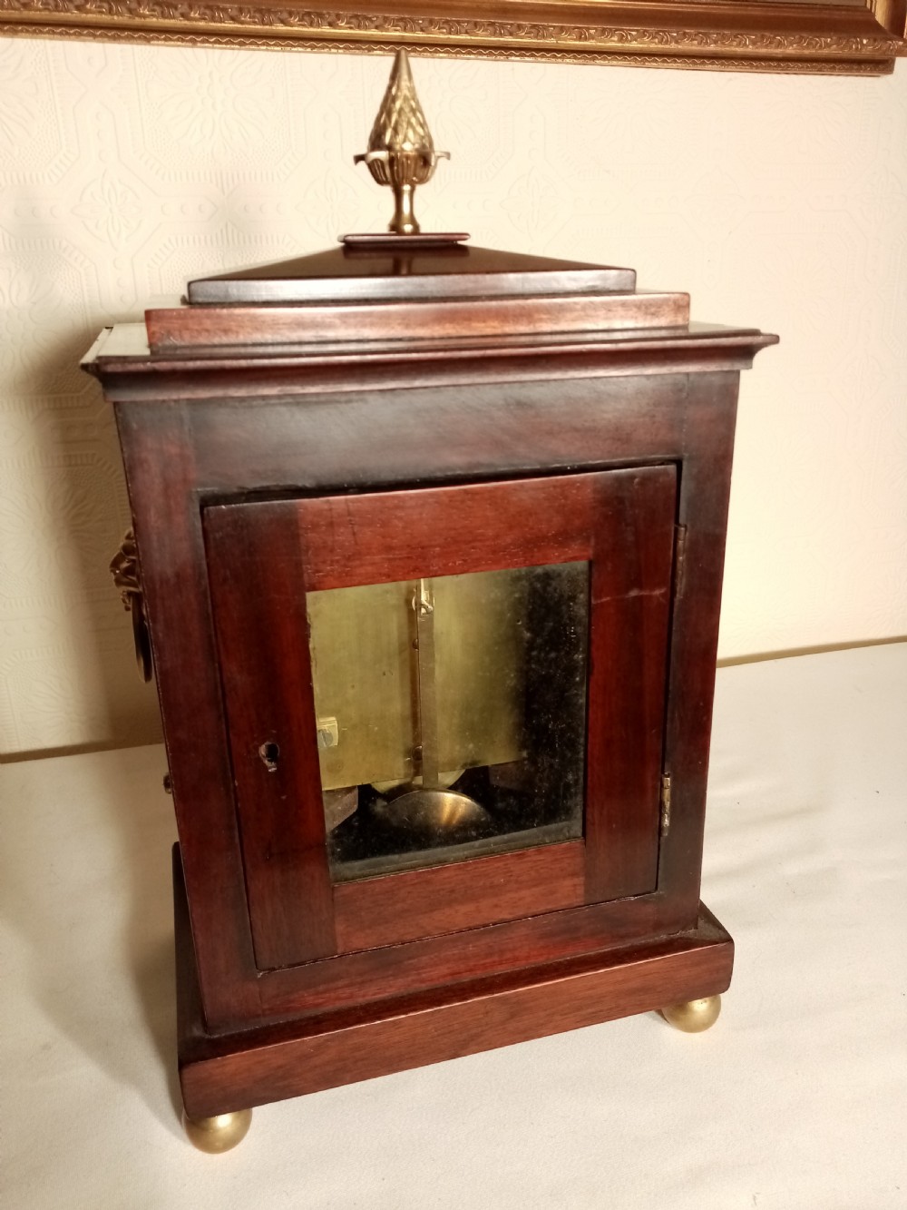 A Regency period dark mahogany library clock featuring brass inlays, a round white dial with Roman numerals, brass ball feet, and a flame finial. View VII