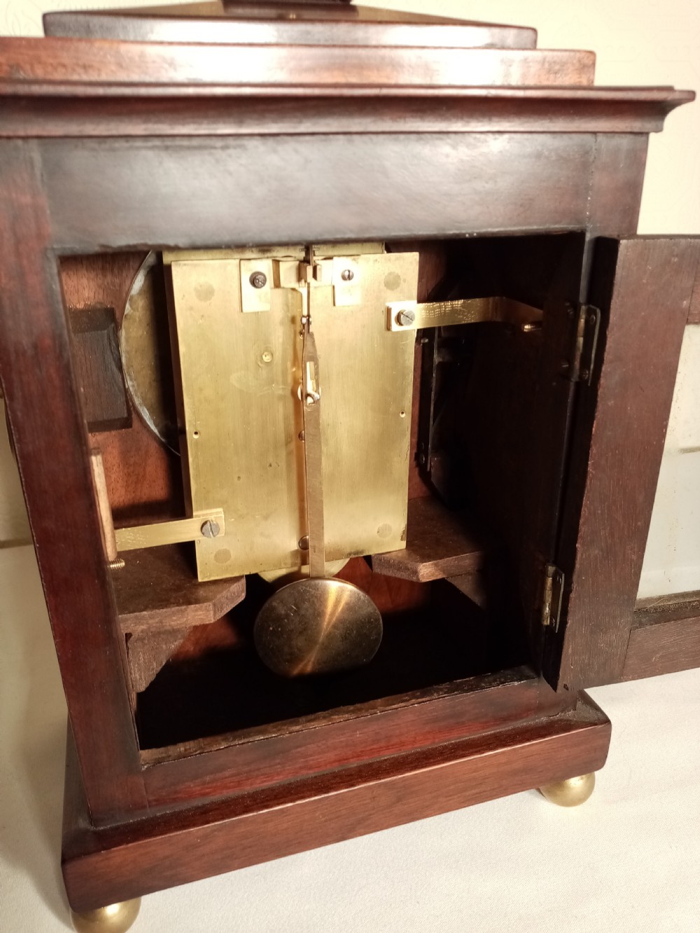 A Regency period dark mahogany library clock featuring brass inlays, a round white dial with Roman numerals, brass ball feet, and a flame finial. View VIII