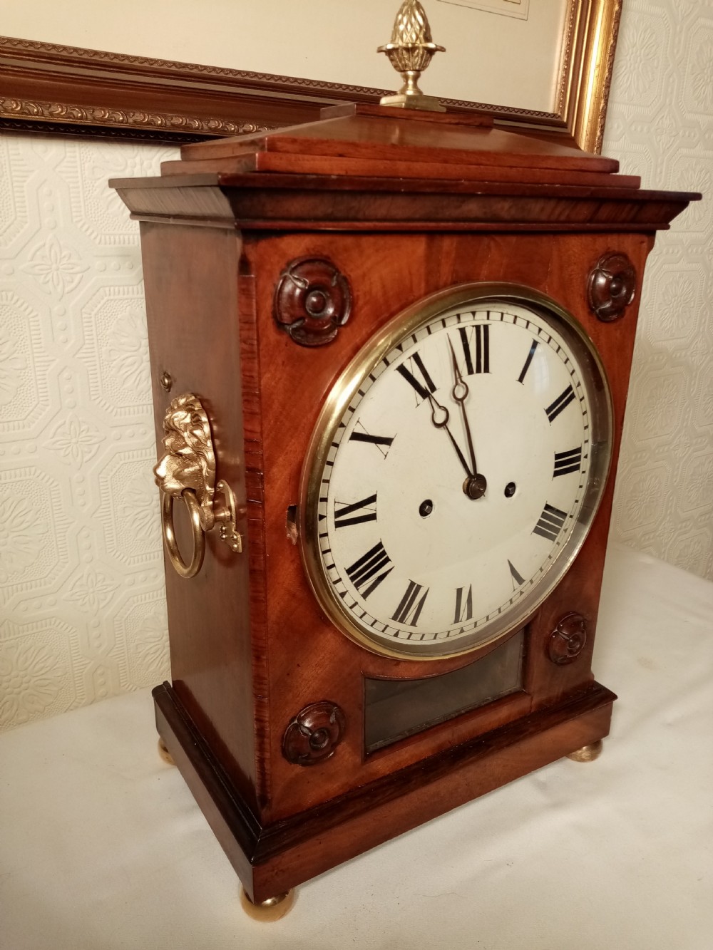 A Regency mahogany bracket clock featuring a circular white dial with Roman numerals, brass lion-head handles, a brass pineapple finial, and a lower glass aperture showing a visible pendulum.
