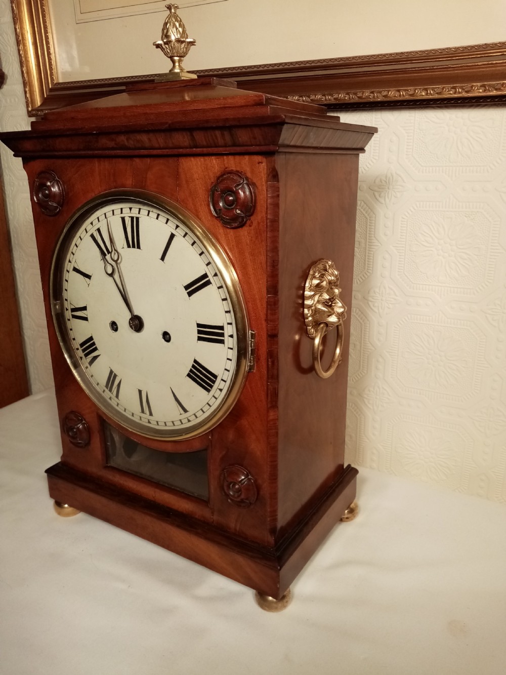 A Regency mahogany bracket clock featuring a circular white dial with Roman numerals, brass lion-head handles, a brass pineapple finial, and a lower glass aperture showing a visible pendulum. View II