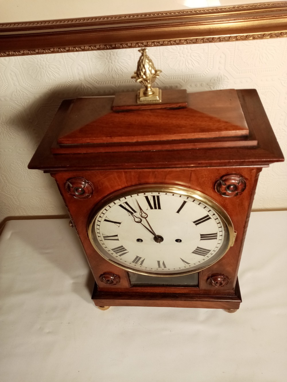 A Regency mahogany bracket clock featuring a circular white dial with Roman numerals, brass lion-head handles, a brass pineapple finial, and a lower glass aperture showing a visible pendulum. View III