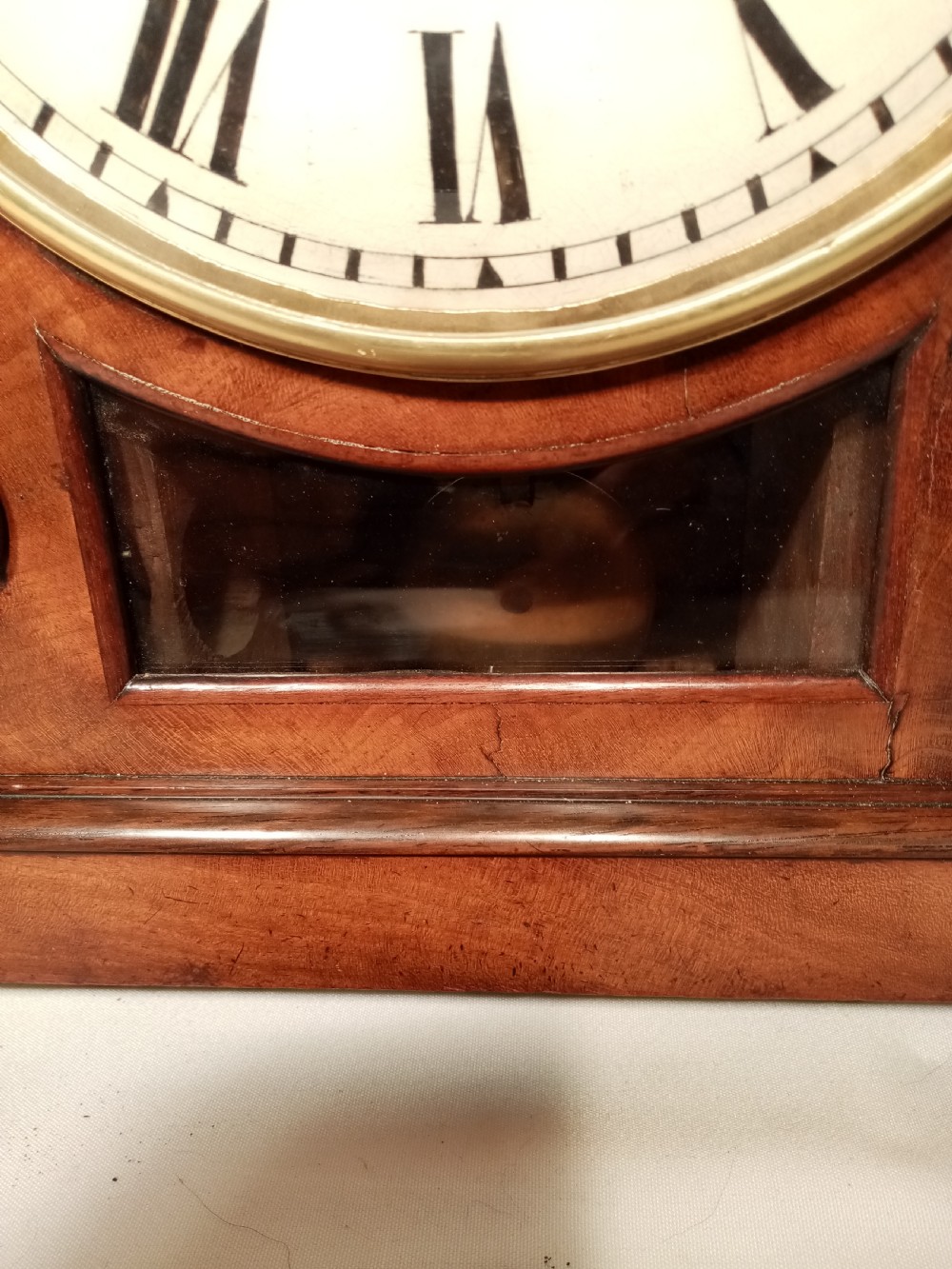 A Regency mahogany bracket clock featuring a circular white dial with Roman numerals, brass lion-head handles, a brass pineapple finial, and a lower glass aperture showing a visible pendulum. View V