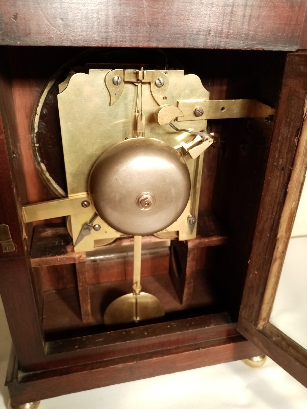 A Regency mahogany bracket clock featuring a circular white dial with Roman numerals, brass lion-head handles, a brass pineapple finial, and a lower glass aperture showing a visible pendulum. View VI