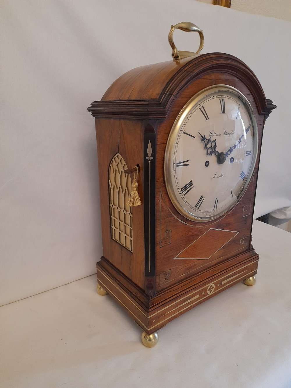 A Regency period rosewood bracket clock featuring geometric brass inlay, arched top with brass handle, gothic side frets, circular dial, and brass ball feet.