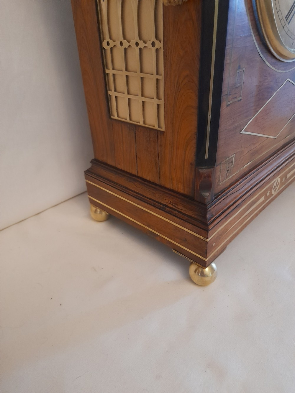 A Regency period rosewood bracket clock featuring geometric brass inlay, arched top with brass handle, gothic side frets, circular dial, and brass ball feet. View VI