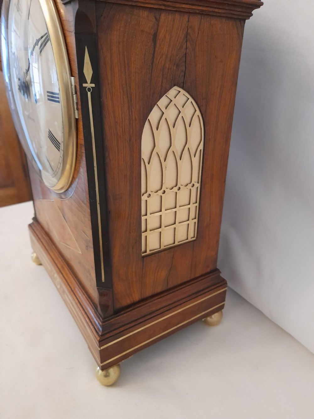 A Regency period rosewood bracket clock featuring geometric brass inlay, arched top with brass handle, gothic side frets, circular dial, and brass ball feet. View VII